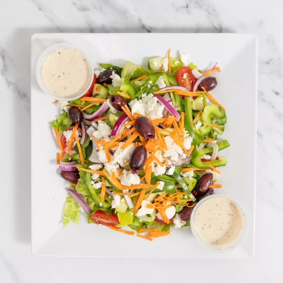 A fresh Greek salad with lettuce, tomatoes, olives, feta, and dressing on a white plate.
