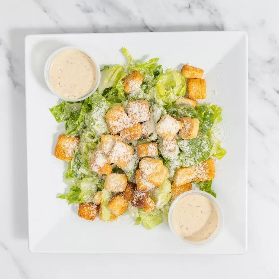 Caesar salad with romaine lettuce, croutons, parmesan cheese, and two cups of dressing on a white plate.