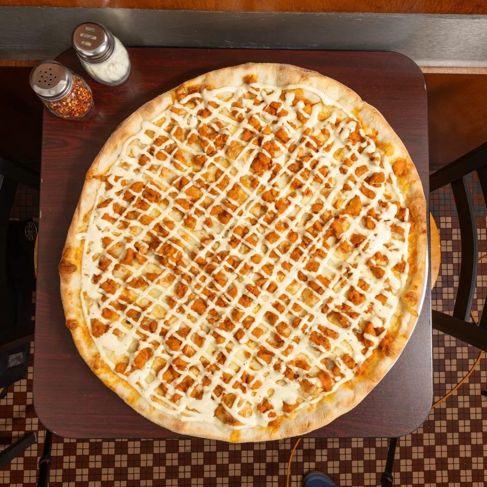 Overhead view of a large buffalo chicken pizza with white sauce drizzle on a dark wooden table.
