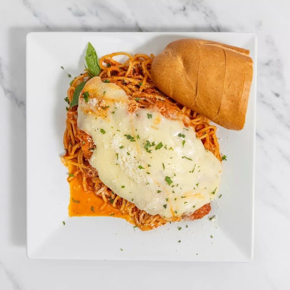 Chicken parmesan with melted cheese, spaghetti, and a side of bread on a white plate.